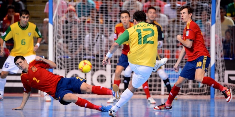 A futsal player kicks the ball towards the opponent's goal while other players try to intercept it.