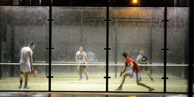 Four athletes play paddle tennis at night on an illuminated court.